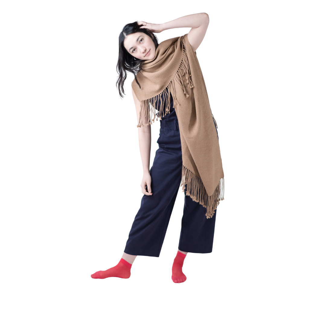 Woman in navy trousers and bright red socks striking a playful pose in a tan alpaca scarf, styled asymmetrically with visible fringe detail.