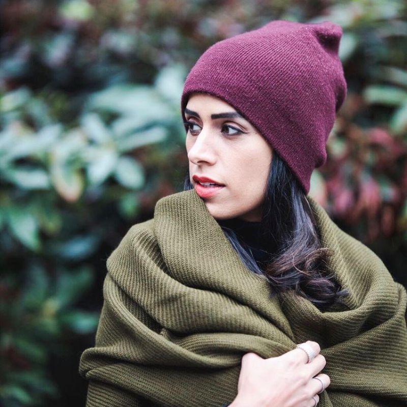 Close-up of a woman, Sonia Beeksma, in a moss green alpaca scarf and burgundy knit beanie, standing in front of dark green foliage and looking to the side.