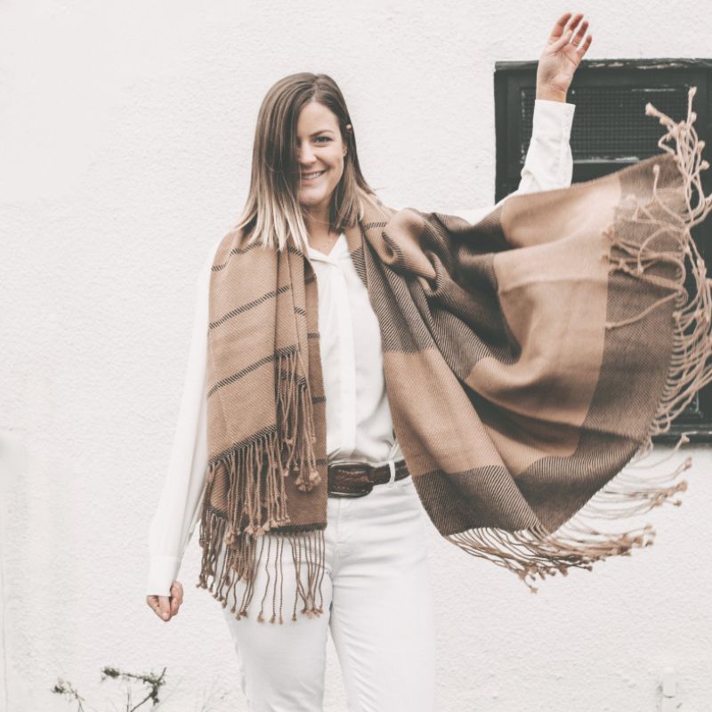 Smiling woman in a white outfit playfully sweeping a camel and black striped alpaca scarf across her body, posed against a white wall.