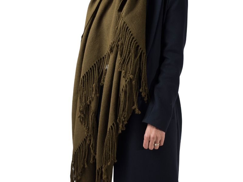 Close-up of a handwoven alpaca scarf in moss green, draped over a dark coat to show long twisted fringe and soft, structured drape.