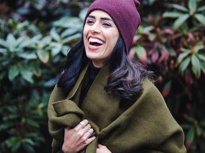 Woman named Sonia Beeksma, host of eTalk, smiling outdoors wearing a moss green alpaca scarf and a burgundy knit hat, wrapped warmly around her shoulders in a natural setting.