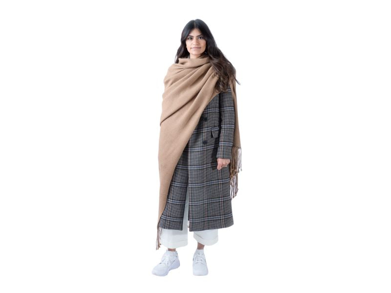 Woman wearing a camel alpaca scarf draped over a long plaid coat and white cropped pants, standing against a white background.