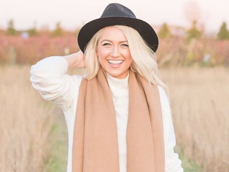 Smiling woman outdoors wearing a camel alpaca scarf over a white sweater and black hat, standing in a golden field at sunset.