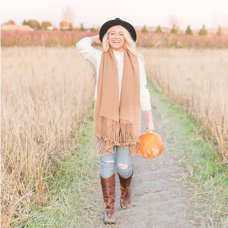 Woman walking through a golden field holding a pumpkin, wearing a camel alpaca scarf with long fringe, paired with a black hat and brown boots.