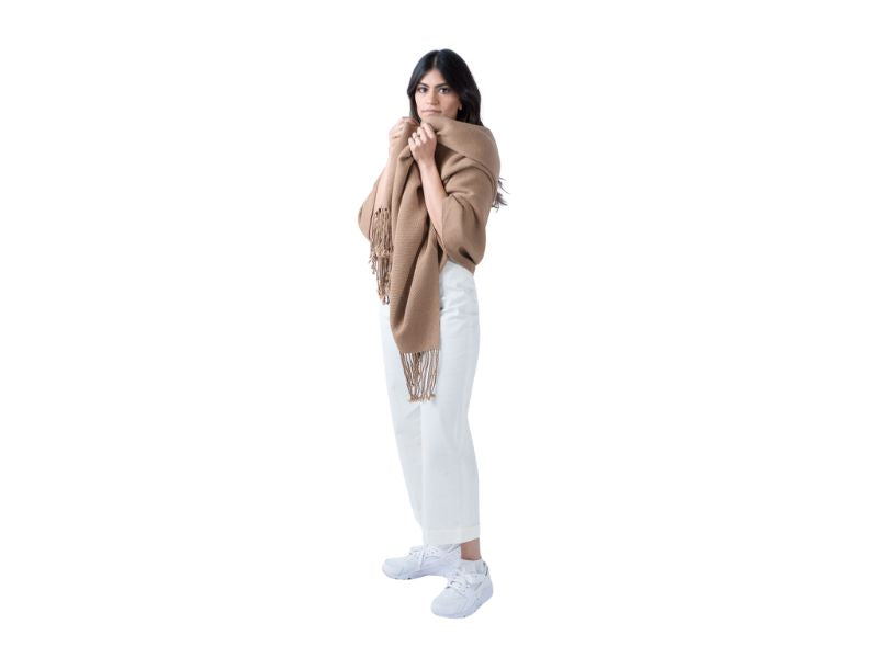 Woman styled in a white outfit with a camel alpaca scarf wrapped over her shoulders, holding the ends close to her face.