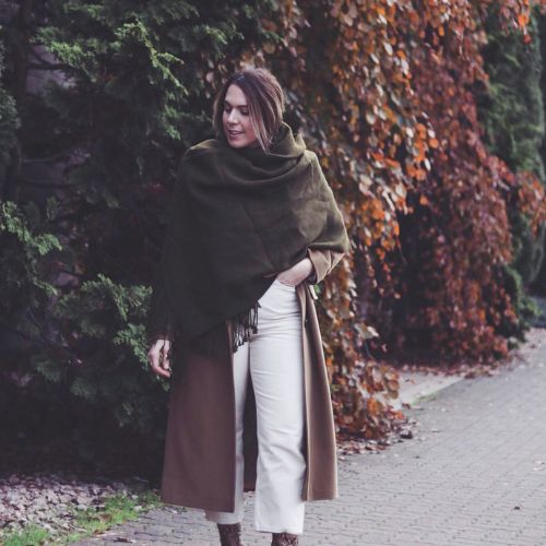 Woman walking on a leaf-lined path in a moss green alpaca scarf and brown coat, surrounded by evergreens and rust-colored ivy.