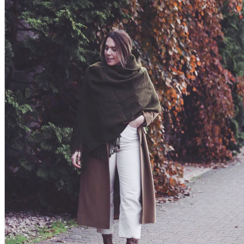 Woman wrapped in a moss green alpaca scarf over a brown coat and white trousers, walking along a tree-lined path with autumn leaves in the background.