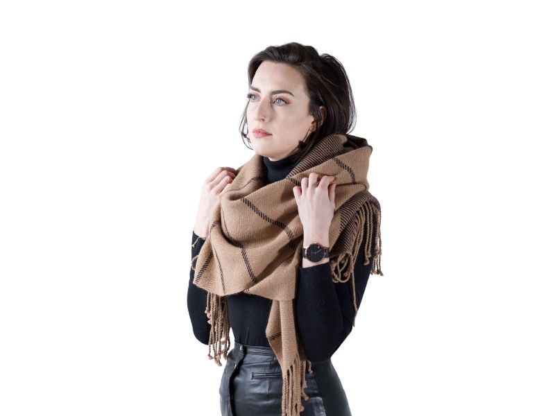 Woman wearing a striped alpaca scarf in camel and black, wrapped high around the neck over a black turtleneck, with a calm studio backdrop.