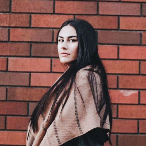 Woman standing in front of a red brick wall wearing a camel alpaca scarf with subtle black stripes, draped softly across her shoulders.