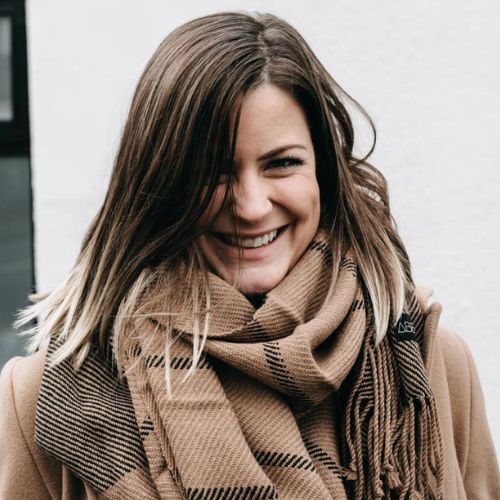 Close-up of a woman smiling in a camel and black striped alpaca scarf, styled over a tan coat against a light wall background.