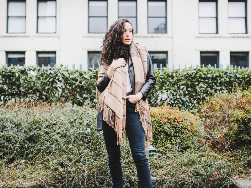 Woman standing in an urban garden wearing a handwoven alpaca scarf with black stripes, styled over a leather jacket and dark jeans.